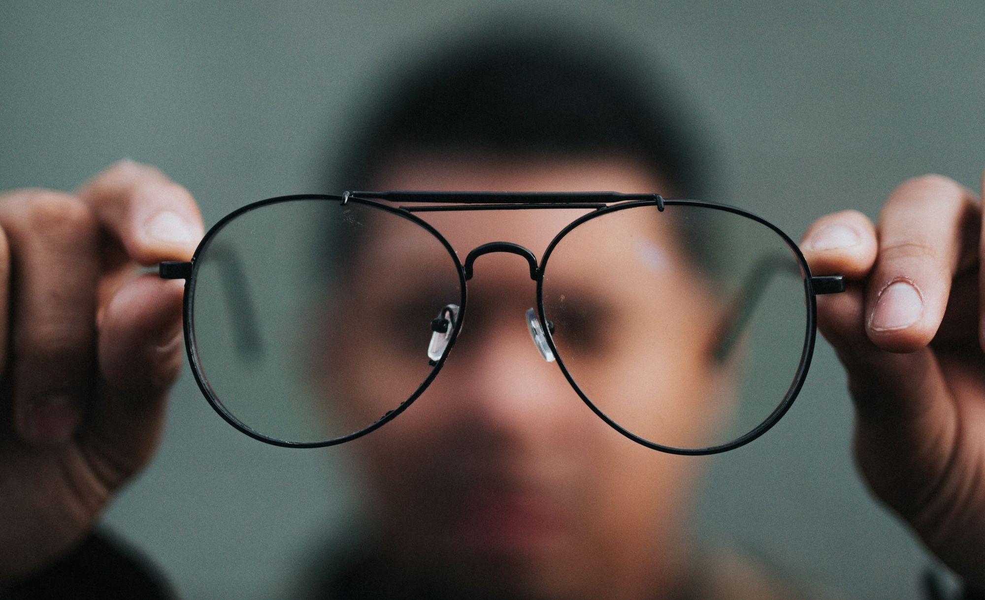 A pair of thin black-framed glasses is in focus, while the man holding them is blurred in the background