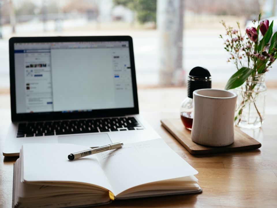 Picture of notebook with a pen balanced on top in front of a laptop. A white coffee mug is set to the right hand side.